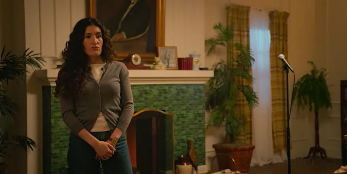 Kate Berlant as Riley standing in her living room beside a lone microphone stand