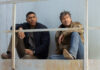 "Lanterns" Aaron Pierre and Kyle Chandler sit on a metal railing, looking pensively into the distance against a weathered backdrop.
