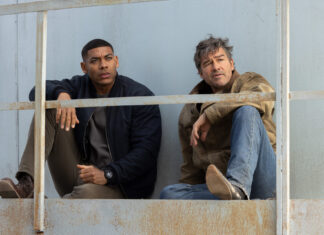 HBO’s “Lanterns” Review: The Green Lantern Series Gets Much Better "Lanterns" Aaron Pierre and Kyle Chandler sit on a metal railing, looking pensively into the distance against a weathered backdrop.