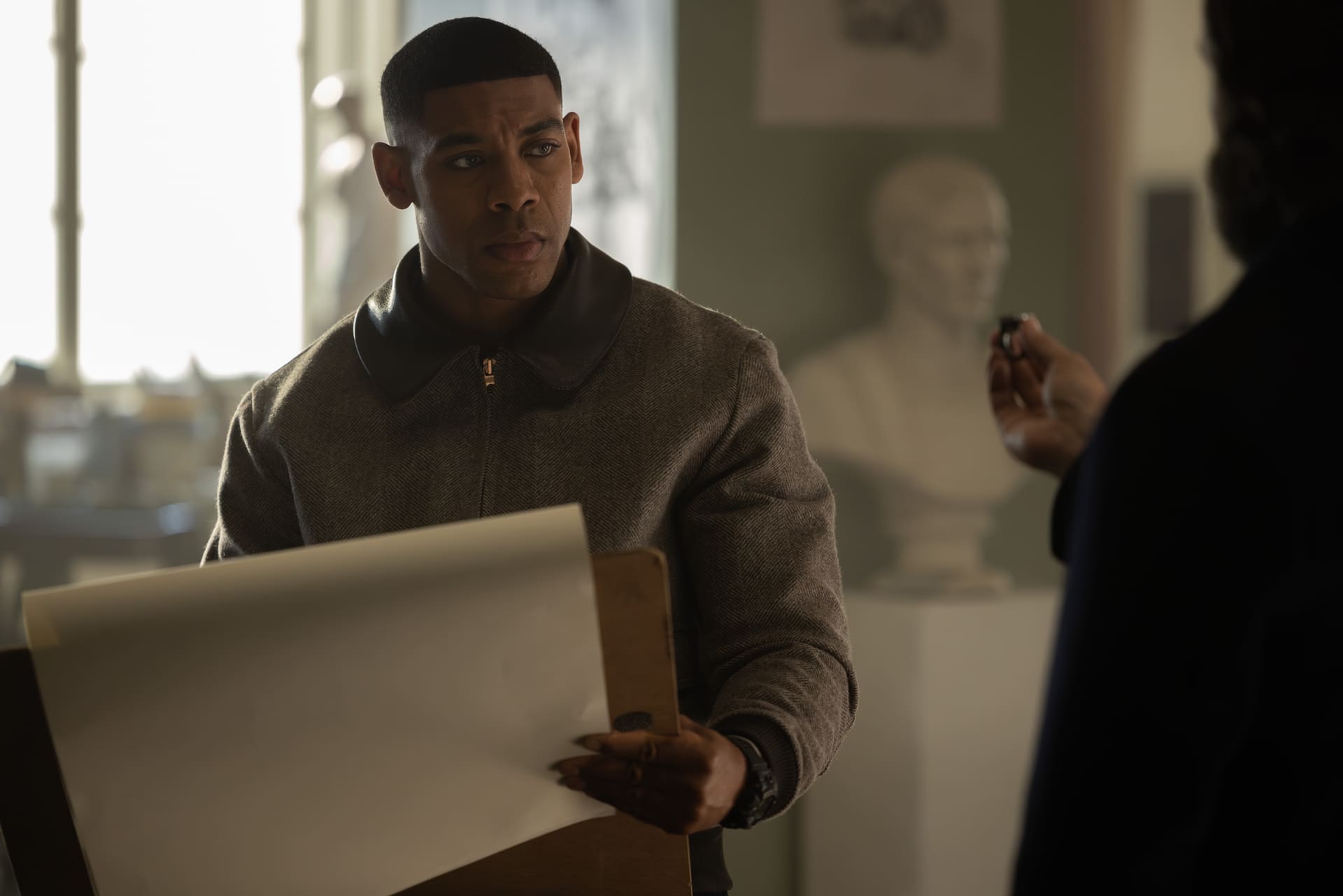Aaron Pierre from "Lanterns," wearing a sweater, holds a drawing pad and looks intently, while someone off-camera appears to hand him something.