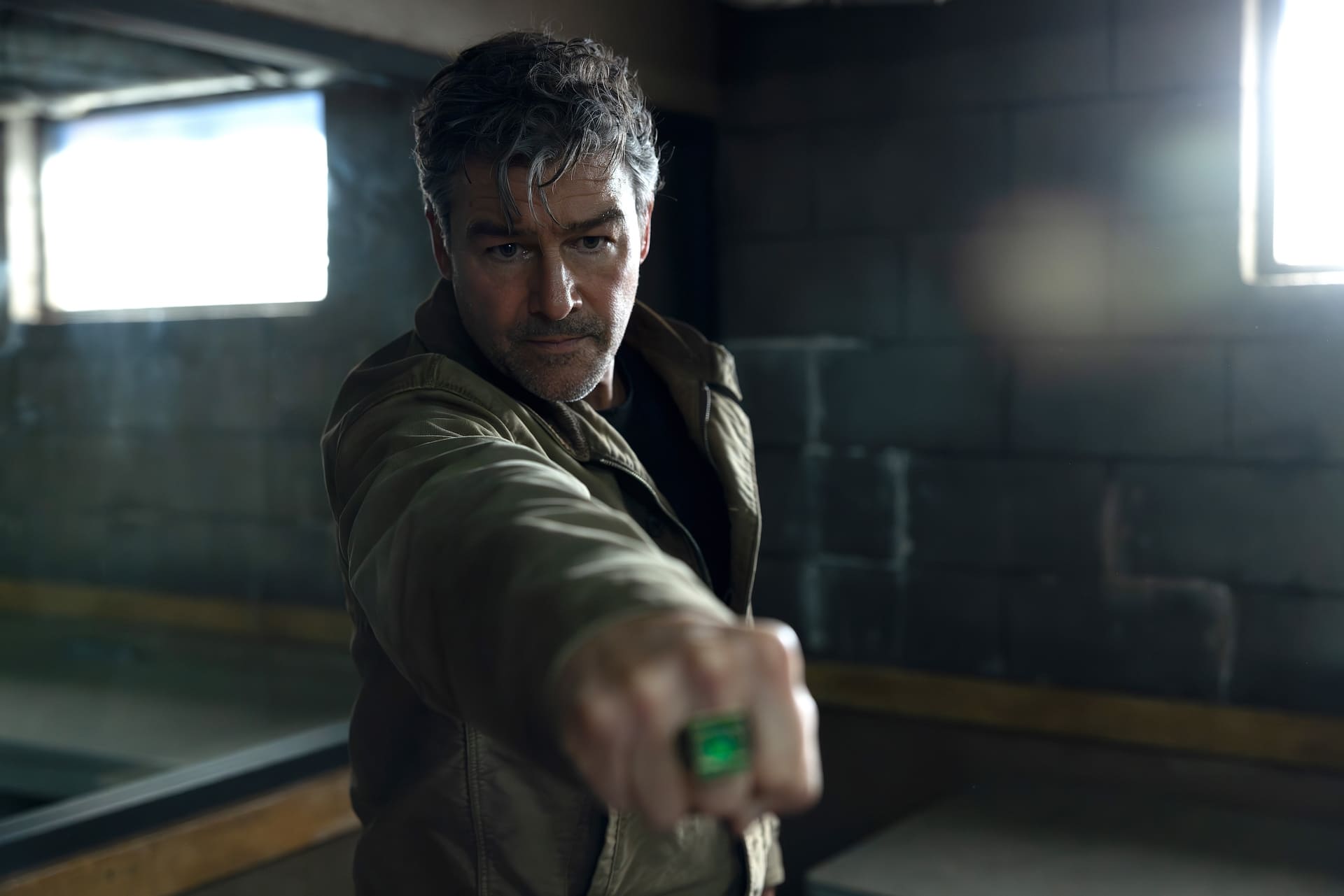 Kyle Chandler from "Lanterns," with short, dark hair and a rugged appearance, is standing in a dimly lit room, wearing a brown jacket and extending his fist forward, displaying a green ring.