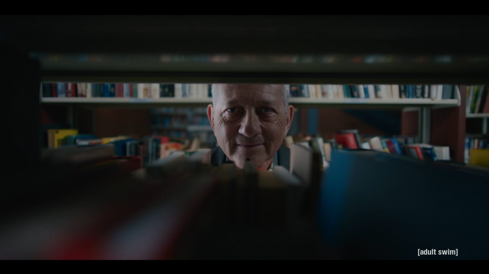 A middle-aged man smiling at Jordan Mendoza through a gap in library shelves.