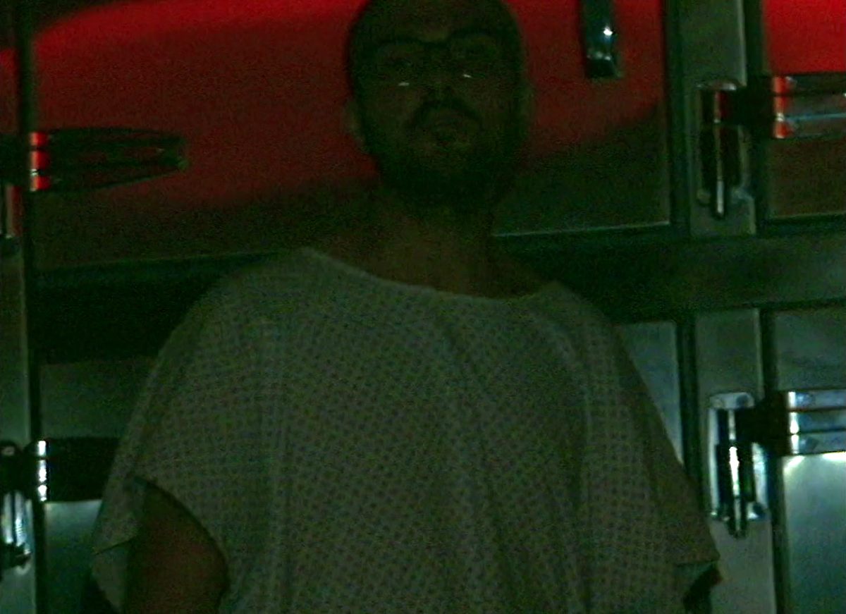 A patient in a hospital gown in a dark corridor, talking straight into the camera